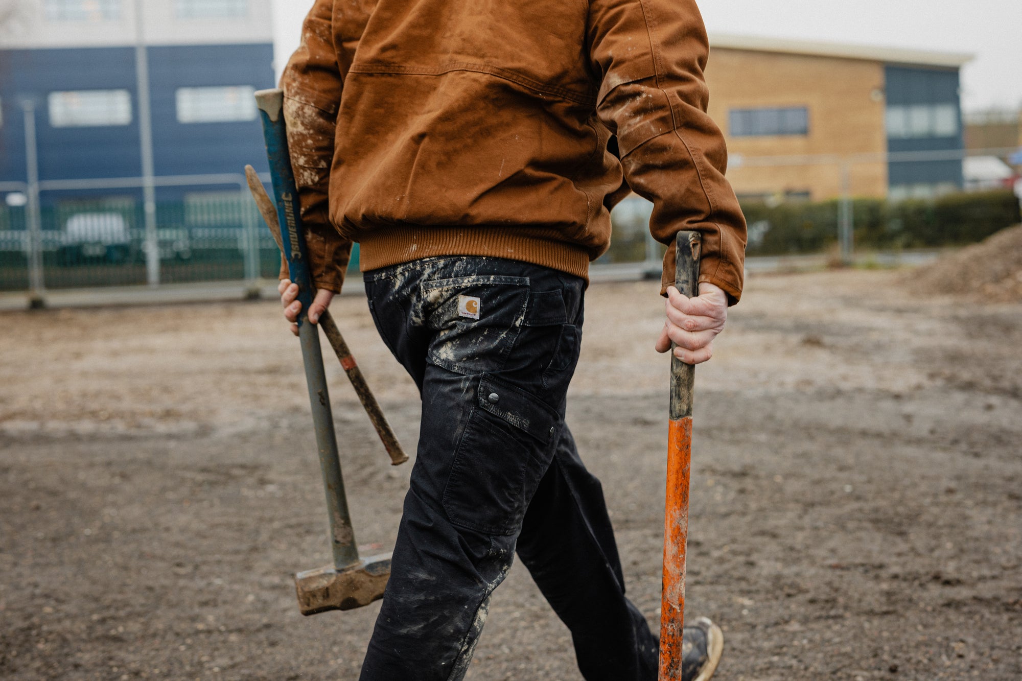 A person from behind wearing a brown Carhartt jacket and dark work pants covered in white paint, holding a sledgehammer and an orange-handled tool on a dirt ground with buildings in the background.